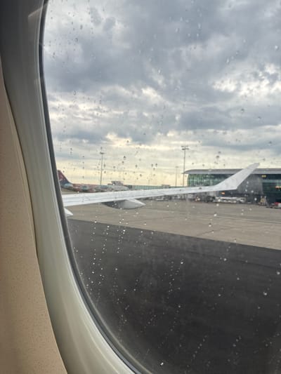 The image is a view from an airplane window, looking out at an airport tarmac under a cloudy sky. Raindrops are visible on the windowpane. In the distance, an airplane wing is prominent, and another airplane with a colorful livery is parked on the tarmac. Airport buildings and structures, including terminals and lighting poles, are visible in the background. The overall scene suggests the view is from Brussels Airport, located in Steenokkerzeel, Belgium, during inclement weather.