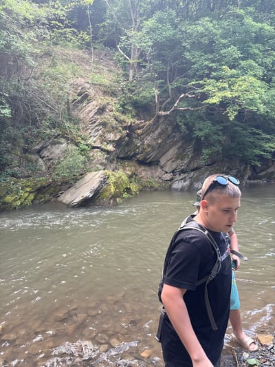 A young male, wearing a black t-shirt, a backpack, and sunglasses perched on his head, stands in shallow water by the bank of a river. His skin tone is fair, and he has short, light-colored hair. Behind him, another person, partially visible, wears a patterned hat and a backpack. The river flows gently, with rocks visible beneath the surface and along the shore. The background features a steep, rocky incline covered with dense green foliage and trees, characteristic of a natural, forested environment. The lighting suggests an overcast day. This scene appears to be in a natural setting, possibly a hiking trail or a recreational area near Lukovo, Bulgaria, given the typical landscape.