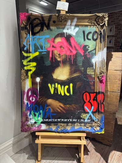 The image presents a vandalized print of the Mona Lisa displayed on an easel. The print is defaced with colorful graffiti, including words like "ICON," "LISA," and "VINCI." The ornate frame surrounding the print is also adorned with graffiti. The overall style suggests contemporary street art or a commentary on high and low culture. The OCR mentions "Ghost Modern Lisa Limited Edition £1695" along with "I AWOKE ONLY TO SEE THAT THE REST OF THE WORLD IS STILL ASLEEP" is an inscription on the art itself. No people are visible in the image. The location, Windermere, United Kingdom, is not visually verifiable in this image.