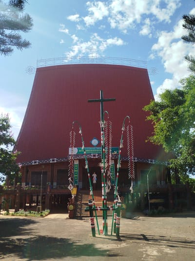 The image captures a unique architectural structure, seemingly a church, in An Phu Ward, Vietnam. The building features a distinctive, steep-sloped red roof, resembling a tribal longhouse. The roof is topped with a decorative metal structure and a cross. Below the roof, the building has a wooden facade with windows and decorated columns. In front of the building, there is a series of green and red painted bamboo poles with hanging decorations. A central cross is positioned amidst the poles. A paved area extends in front of the building, marked by shadows from nearby trees. A few people are visible near the entrance of the church. The sky is blue with scattered clouds. The overall environment appears to be lush with greenery.