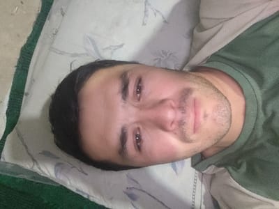A single adult male is depicted in an indoor, close-up, chest-up shot. He is lying on his back, facing the camera directly, with his head resting on a light-colored pillow featuring a subtle, muted blue/grey floral or leaf pattern. The subject appears to be in his 20s or 30s, with short dark hair, dark eyes, and visible facial stubble forming a short beard and mustache. His expression is neutral. He is wearing a dark green, crew-neck shirt. A lighter, possibly beige or off-white, striped fabric is visible beneath his right shoulder and arm, consistent with bedding material. To the left of the pillow, a dark green, textured mat or carpet is partially visible. The lighting is uniform, suggesting an interior space. The image provides no discernible visual information to confirm the location as Yangikurgan, Uzbekistan.