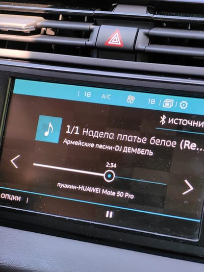 The image shows a close-up of a car's infotainment system screen displaying music playback information. The screen is predominantly black with teal and white text and icons. At the top, there are indicators for "A/C", fan speed, and possibly battery life or signal strength. The central part of the screen shows a music track titled "1/1 Надела платье белое (Re..." which translates to "1/1 Wore a white dress (Re...". Below this, the artist is listed as "Армейские песни-DJ ДЕМБЕЛЬ" meaning "Army songs-DJ DEMBEL". A music note icon is visible on the left. A progress bar shows the current playback time as "2:34". The song title or artist also appears to be "пушкин-HUAWEI Mate 50 Pro" further down. There are navigation arrows on the left and right sides of the screen. At the bottom left, the word "ОПЦИИ" (OPTIONS) is visible. The environment is dark, suggesting the interior of a car, and the lighting indicates the screen is illuminated. The background shows parts of the car's dashboard, including air vents and a hazard light button. The image's metadata does not contain information about the location in the city of Ilyanskiy selskiy Sovet, Belarus.
