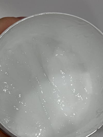 The image shows a close-up, top-down view of a white, viscous substance inside a silver-colored container. The substance appears to be a cream or gel, with some sheen and visible texture from swirling. A human finger is partially visible on the left side, indicating the container is being held. The background is a plain, light gray. There are no discernible landmarks or environmental features to suggest a specific location within Likino-Dulyovo, Russia.