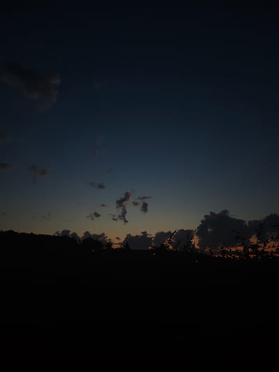 The image depicts a twilight sky over a silhouetted landscape, likely taken in Wilnsdorf, Germany. The sky transitions from a deep navy blue at the top to lighter shades of blue and orange near the horizon. Scattered clouds are visible, some dark and appearing textured, while others are wispy and less defined.
The lower portion of the image is dominated by the dark silhouette of a tree line or a low hill, with indistinct shapes that could be trees or bushes. A few small, faint lights are visible against the dark silhouette, possibly streetlights or distant homes. The foreground is entirely black, suggesting it is either very dark or cropped to exclude detail. The overall impression is of a peaceful, natural scene at dusk or dawn.