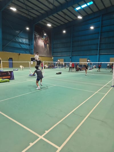 This is a photograph taken at the Shri Guru Gobind Singh Stadium in Phagwara, India, a multi-purpose indoor sports complex known for hosting badminton tournaments. The image captures a badminton match in progress on a green court with white lines.
In the foreground, a young male badminton player, wearing a dark grey t-shirt with "SINGH.B" printed on the back and blue shorts, is in the middle of a shot, his body twisted and racquet extended. He is on the left side of the court.
Behind him, on a raised umpire's chair, a male umpire in a white shirt and black pants is observing the game. A score marker with the number '5' is visible on the chair.
Several other individuals are present on the court and in the background. On the right side of the net, there are players, spectators, and officials. Some are standing near the net, while others are seated. A large banner with a picture of Saina Nehwal, a renowned Indian badminton player, is displayed on the back wall.
The stadium's interior features a high ceiling with exposed metal beams and lighting fixtures. The walls are painted in a blue hue, and the overall atmosphere suggests a competitive sporting event. The lighting is a mix of natural light from windows at the top and artificial illumination from overhead lamps.