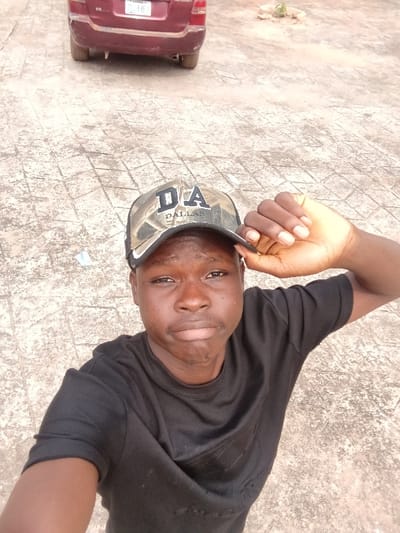 A young male with dark skin is depicted in a high-angle selfie. He wears a black short-sleeved t-shirt and a camouflage-patterned baseball cap with "DA" prominently displayed and "DALLAS" written beneath it on the front. His right hand is raised, with fingers resting on the brim of the cap. The male's facial expression is neutral. In the upper background, a maroon-colored vehicle, possibly an SUV or sedan, is partially visible, facing away from the camera. The ground is a light-colored, patterned paved surface, consistent with interlocking concrete pavers. A small circular area of disturbed earth or sparse vegetation is visible in the upper right background. The scene is outdoors, in the city of Itu, Nigeria.