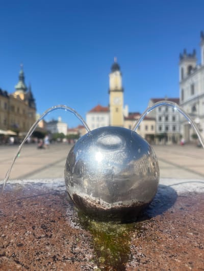 The image depicts a close-up of a spherical fountain element made of polished metal, with two streams of water arching upwards from its surface. The fountain is situated on a textured, reddish-brown surface, possibly granite, which appears wet around the base of the sphere, with some green algae visible. In the background, a city square is visible under a clear blue sky. Buildings with historical architecture, including a tall clock tower with a yellow facade and a white multi-story building with decorative features, are blurred due to the shallow depth of field. A few indistinct figures of people can be seen walking in the distance. This scene is located in the SNP Square (Námestie SNP) in Banská Bystrica, Slovakia, a central public space often featuring fountains and surrounded by historical buildings.