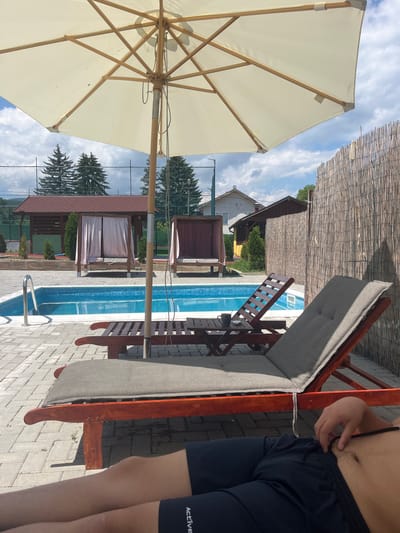 The image depicts an outdoor recreational area in Bersin, Bulgaria, under a partially cloudy daytime sky. In the immediate foreground, a person is reclining on a wooden lounge chair with a grey padded cushion. Only the lower torso and legs of the person are visible, dressed in dark blue or black shorts with "ACTIVE" text on one leg; one hand is resting near the waist.
To the left of the reclining person, another similar wooden lounge chair is visible with its backrest in an upright position. A small, dark wooden side table is situated between these two chairs, holding a dark-colored mug or cup. A large, cream-colored patio umbrella with a wooden frame shades this foreground area, its central pole and internal structure clearly visible.
Beyond the lounge chairs and umbrella, a rectangular swimming pool with blue mosaic tiles is visible, featuring a stainless steel handrail at one end. Further in the background, past the pool, are two open-sided wooden gazebos or cabanas with draped curtains; the left one has light-colored curtains, and the right one has darker, possibly reddish-brown, curtains. Behind these structures, a line of green coniferous trees is present, and a green mesh fence stretches horizontally across the mid-ground. To the far right, a tall fence constructed from natural reed or bamboo material runs vertically along the edge of the scene. In the distance, behind the trees and fences, parts of residential-style buildings, including one with a white facade and another with a yellow/brown facade, are discernible under a sky with scattered white cumulus clouds.