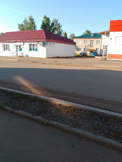 The image captures a street scene in Kranokholmskiy, Russia, under a clear blue sky. In the foreground, a gray asphalt road with a concrete curb and a narrow strip of gravel and sparse vegetation runs across the bottom of the frame. A long shadow stretches across the road from the left.
In the middle ground, a white building with a red tiled roof, labeled "АПТЕКА" (Pharmacy), is visible on the left. It has several windows and an air conditioning unit. Next to it, to the right, are two other buildings. One is a lighter-colored, multi-story building with a green metal roof and multiple windows. In front of this building, two cars are parked: a dark-colored sedan and a small blue hatchback. To the far right of the image is a white building with a red roof and brown-colored siding. A sign with the number "2" is visible on this building. The background consists of a line of green trees.