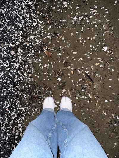 The image shows a close-up, high-angle view of a person's legs and feet standing on a dark, sandy beach. The person is wearing light blue denim jeans and white sneakers. The sand is speckled with numerous small white and dark shell fragments, as well as some dried seaweed and debris, including a single black feather. In the upper left portion of the frame, the sand is darker and less populated with shells, while the lower portion of the frame is heavily covered with a dense layer of white shell fragments. The lighting suggests it might be dusk or nighttime, as the overall illumination is low. The scene is a typical beach environment. There is no visible text in the image. The context provided states the location is Chiba, Japan.