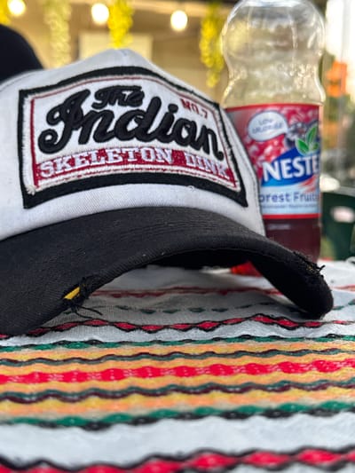 The image shows a close-up of a black baseball cap and a bottle of Nestea iced tea. The cap has a white patch with black embroidered text that reads "The Indian Skeleton Crew". The bottle of Nestea is mostly out of focus but its label is visible, showing the brand name "NESTE" in blue letters and "Forest Fruits" in red. The background is blurred, with hints of greenery and yellow lights, suggesting an outdoor setting, possibly during the day. The fabric to the right of the cap has a colorful striped pattern with wavy lines in red, yellow, green, and black. A small yellow object is visible on the black fabric of the cap. The image is oriented vertically.