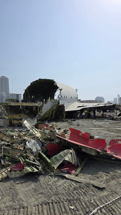 The image depicts the wreckage of a large aircraft, possibly a Boeing 747, scattered across a dry, dusty lot in Fujairah, United Arab Emirates. The main fuselage section is heavily damaged, with a large portion of the upper hull torn away, revealing the internal structural framework. Sections of the plane's exterior, including large white and red panels, are strewn about the foreground and midground, intermingled with debris. Several figures, presumably workers wearing high-visibility vests, are visible near the wreckage, suggesting ongoing salvage or dismantling operations. In the background, modern high-rise buildings of Fujairah can be seen against a clear, pale blue sky, providing a stark contrast between the urban development and the scene of destruction. The ground is a mixture of dirt and gravel, marked with tire tracks and footprints.