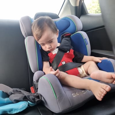 A toddler is asleep in a car seat, which is positioned on the back seat of a car. The car seat is gray with blue mesh inserts and green trim. The toddler is wearing a black t-shirt with a red insert and yellow striped shorts. The toddler's arms are resting on their lap, and their legs are extended. The car seat is secured with a black harness. In the background, a window of the car shows a blurred view of trees and foliage, suggesting an outdoor setting. On the floor of the car, next to the car seat, is a blue inflatable cushion and a dark gray folded garment. The overall scene is domestic and depicts a child traveling in a vehicle. The location is Bongpyeong-myeon, South Korea.