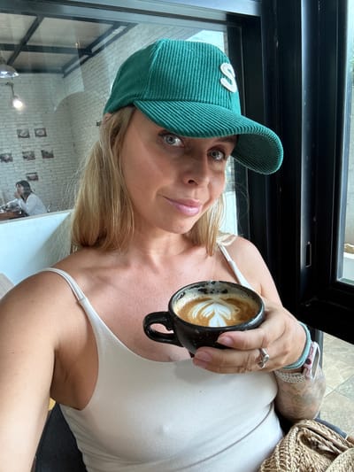 A blonde woman wearing a green corduroy baseball cap with a white "S" on the front and a white tank top holds a black mug of coffee with latte art in her right hand. She is seated indoors in what appears to be a cafe, with a reflection of another woman in the background visible through a window. The environment shows exposed brick walls and pendant lights. The woman has fair skin, blue eyes, and light pink lipstick. She is also wearing rings on her fingers and bracelets on her wrist. A woven bag is visible in the lower right corner of the image. The location is not specified as Tha Chana, Thailand.
