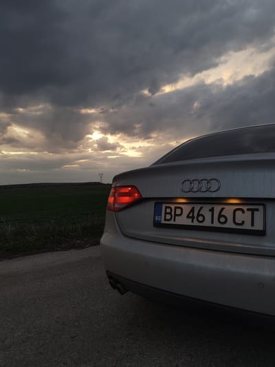A silver Audi sedan, displaying Bulgarian license plate 'BP 4616 CT', is parked on the asphalt shoulder of a road in Gorni Tsibar, Bulgaria. The vehicle is positioned facing away from the viewer, with its rear-left quarter visible. The driver-side taillight is illuminated red, and the license plate lights are active, casting an amber glow. Dual exhaust pipes are visible on the lower left rear, and circular parking sensors are embedded in the bumper. To the left of the road, a green field extends into the distance. A tall, lattice-style metal power line tower is visible on the horizon, silhouetted against the sky. The sky is dominated by dark, heavy grey clouds, through which distinct patches of golden-orange light are breaking, suggesting either sunset or sunrise. No individuals or direct actions are observable.