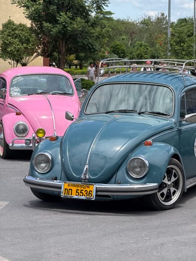 The image shows two Volkswagen Beetles parked next to each other. The front Beetle is a bright pink and the one behind it is a teal blue. Both cars appear to be older models, likely from the 1960s or 1970s. The pink Beetle has a chrome bumper and a yellow headlight. The teal Beetle has a chrome roof rack and a yellow license plate with Thai characters and the number "5536". The cars are parked on a paved surface, possibly a street or parking lot. In the background, there are trees and some people can be seen through the windows of the teal Beetle. The lighting suggests it is daytime, and the weather appears to be clear. The overall impression is that of a car meet or a casual gathering of car enthusiasts. The image is taken from a slightly elevated angle, looking down on the cars. The scene is static, with no apparent activity taking place. The cars are the primary subjects of the image.