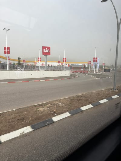 The image captures an outdoor commercial area, likely a shopping center or retail park, viewed from a road on an overcast or hazy daytime. In the foreground, a paved road with black and white striped curbs runs along the right side, adjacent to a strip of dirt. To the left, a concrete barrier with red and white striped markings separates the road from a large retail complex.

Numerous tall white poles, each topped with a prominent red square sign displaying the word "BIG" in white, are visible across the scene, indicating the name of the shopping center. Several Israeli flags, featuring a blue Star of David on a white background, are also flown from similar poles, particularly in the mid-ground.

Beyond the concrete barrier and signs, various low-rise commercial buildings can be seen. A distinctive yellow and red McDonald's restaurant is identifiable, with its logo and name clearly visible. Other building facades bear partial brand names like "FOX HOME" and "Golf," along with Hebrew text, suggesting a range of stores. A small tree stands amidst the buildings in the distance. The sky is uniformly light gray, indicating either heavy cloud cover or atmospheric haze, with no direct sunlight. No people are visible, and no specific activity is taking place; the scene is static, showcasing the infrastructure of the commercial district in Migdal HaEmek, Israel.