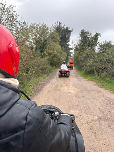 The image presents a first-person view from a quad bike navigating a muddy dirt track in Çiftlikköy, Turkey. In the immediate foreground, the left shoulder and arm of an individual wearing a black jacket are visible, along with a portion of a red helmet on the left side. The individual's hands are gripping the handlebars of a black quad bike. Ahead, two additional quad bikes proceed in a single file along the unpaved track, which features multiple visible puddles. The first quad bike in the line is red, driven by a person wearing a light-colored hooded garment and a white helmet. The second quad bike is dark-colored, operated by an individual dressed in an orange high-visibility jacket and a dark helmet. The track is bordered on both sides by dense vegetation, consisting of trees and shrubs with green and brown foliage. The sky above is uniformly overcast and grey.