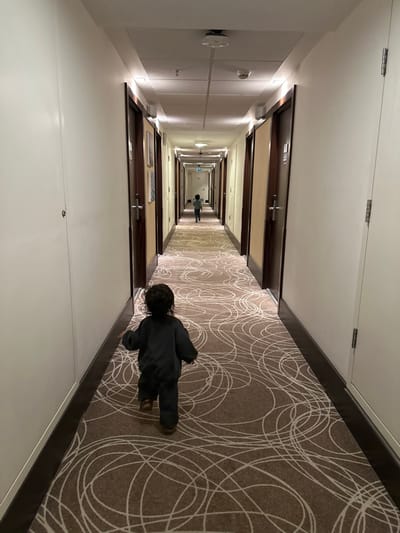 The image displays a long, well-lit interior hallway within a building in Zeytinburnu, Turkey. The hallway features light-colored walls and is lined on both sides with multiple dark wooden doors set into dark brown frames, each equipped with metal handles. The floor is covered with a brown carpet patterned with large, interlocking off-white circular designs. The white ceiling contains recessed lighting fixtures, several circular white smoke detectors, and a sprinkler head.

In the foreground, a young child with dark, curly hair, dressed in a dark long-sleeved top and dark pants, with visible light-colored footwear, is depicted from behind, walking or running away from the viewer. Further down the hallway, a second, smaller child, also viewed from behind and wearing a light-colored top with dark pants, is moving in the same direction. A green emergency exit sign is visible at the far end of the hallway.