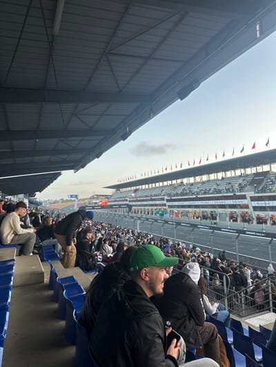 The image displays a crowded grandstand at the Circuit de la Sarthe in Le Mans, France, with a view of the racetrack and pit complex. In the immediate foreground, spectators occupy rows of blue plastic seating and stand on concrete steps beneath a grey corrugated metal roof structure, supported by steel beams and equipped with black speakers. A man wearing a green baseball cap and black jacket, holding a smartphone, is seen in profile, smiling and looking towards the track. Adjacent to him, another person wears a white beanie and a dark jacket. On the far left, a young man in a cream-colored sweater and jeans sits on a concrete ledge.
The midground reveals a dense multitude of spectators filling further grandstand sections, facing a multi-lane asphalt racetrack. Beyond the track, a complex of multi-story buildings, likely comprising pit garages or corporate hospitality suites, features numerous large windows and prominent "SAMSUNG Galaxy A" branding. A long line of national flags on poles adorns the top of these structures. In the background, additional grandstands, also populated with spectators, are visible under a pale blue sky with scattered light clouds.