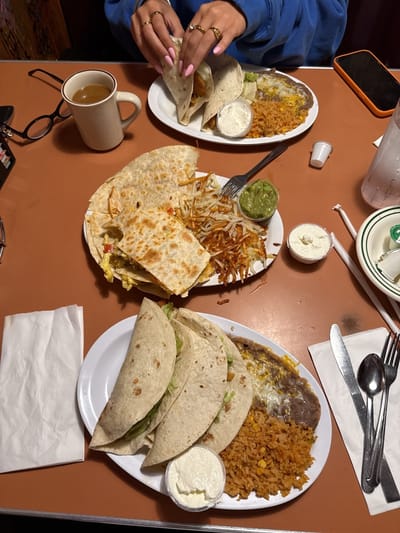 The image depicts a table at a restaurant, likely in Huntington Beach, United States, based on the style of food. Three plates of food dominate the scene. One plate has a quesadilla cut into pieces with a side of hash browns and guacamole. Another plate holds three tacos, rice, refried beans, and a small container of sour cream. The third plate shows two tacos, rice, refried beans, and a small container of sour cream. A person with pink fingernails is holding one of the tacos on the second plate. Other objects on the table include a mug of coffee, eyeglasses, a cell phone, cutlery, a glass of water, a small container of cream, napkins, and condiment containers. The tabletop is orange.