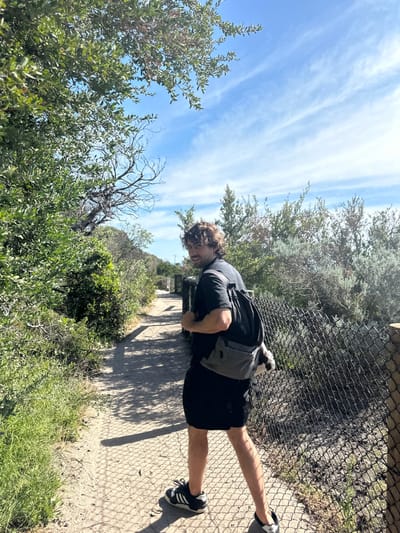 A person stands on a concrete path beside a wire fence in a green, natural setting. The individual is wearing a dark t-shirt, shorts, and black and white sneakers. A light gray backpack is slung over their shoulder. Lush green foliage and trees surround the path, contrasting with the bright blue sky filled with wispy clouds. The perspective of the shot is canted, creating a tilted view of the scene.