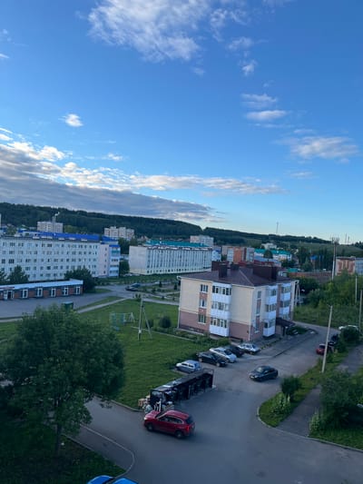 The image depicts a panoramic view of a residential area in Leninogorsk, Russia, under a bright blue sky with scattered clouds. In the foreground, a red SUV is parked on a paved road next to a row of large trash receptacles. Several other cars are parked in a lot to the left, including a blue sedan. A mature tree with lush green foliage dominates the left side of the frame.
In the mid-ground, a multi-story apartment building with a beige and white facade and a brown roof stands prominently. A playground with swings and a climbing structure is visible on a grassy area adjacent to the building. Further back, several other apartment buildings of varying heights and architectural styles are visible, some with blue roofs. The landscape rises behind the buildings, covered in a dense green forest. In the distance, communication towers and industrial chimneys are discernible. The overall scene suggests a typical day in a Russian city, with residential buildings, infrastructure, and natural elements coexisting.