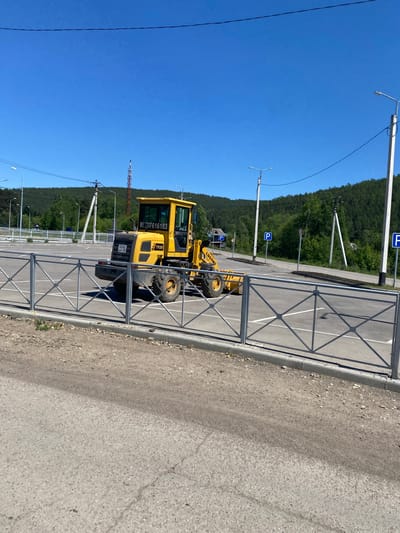 A yellow wheel loader with the brand "LUGONG" and model "T920" is parked in an empty parking lot under a clear blue sky. The vehicle has visible license plates. The parking lot is delineated by white lines and surrounded by a metal fence. In the background, a dense forest covers rolling hills. Several streetlights and utility poles are also present. The scene is set in the city of Bolshoy Lug, Russia.
