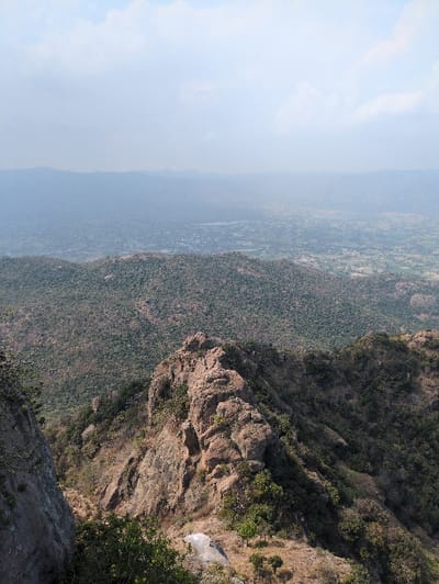 This image depicts a wide panoramic view of a mountainous and valley landscape, likely situated near Kadaladi, India. The foreground features a steep, grey rock face on the left and a prominent, rugged ridge of weathered brown and grey rock formations extending across the mid-right. This ridge is sparsely covered with dry brown grasses and small, green-leaved trees. Beyond this immediate ridge, the midground transitions to undulating hills densely covered in dark green foliage. The background reveals a vast, hazy valley stretching to the horizon, dotted with scattered human settlements and agricultural land. A small town or village with an adjacent body of water is discernible in the distant mid-left section of the valley. The sky is a light, hazy blue with scattered white clouds. No people, specific objects, actions, or interactions are visible within the frame; the scene is purely environmental.