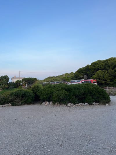 The image depicts an outdoor scene in Sant Pere de Ribes, Spain, characterized by a clear, pale blue sky at what appears to be late day. In the foreground, a light gray gravel path or unpaved road occupies the lower third of the frame. This path is bordered by a scattered line of light-colored rocks, behind which a dense, dark green bush occupies the lower mid-ground.

Beyond the bush, a railway line is visible. A red and white commuter train is moving on these tracks, positioned from the right and partially obscured by the bush. The train's front section is predominantly red, transitioning to white with red detailing along its side. Overhead lines for the train are discernible above the tracks.

To the left of the railway line and behind the bush, a light-colored building, possibly a house, is situated amongst trees. This building has a reddish-brown tiled roof and a tall, slender, metallic structure, resembling an antenna or utility pole, rising from its side. The background is dominated by a densely forested hillside, covered in dark green trees, which slopes upwards from the right towards the center-left. No individuals are present, nor are there any discernible actions or interactions occurring within the frame.