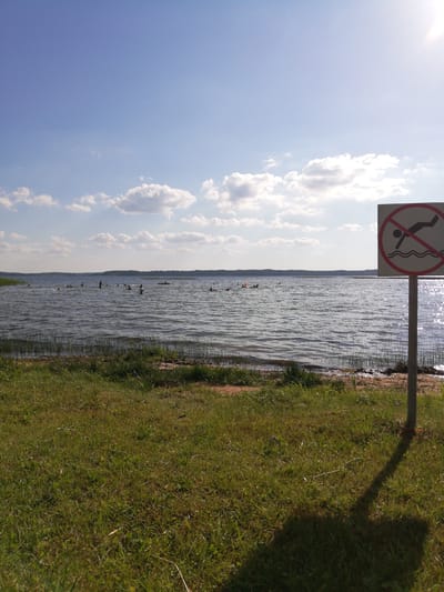 The image depicts a lake with several people swimming or wading in the water. A sign with a red circle and a diagonal line over a silhouette of a person diving is prominently displayed on the right side of the frame, indicating a prohibition against diving. The lake is surrounded by a treeline in the distance, and the foreground features a grassy bank. The sky is blue with scattered white clouds, and the sun is visible, casting a glare on the water's surface. The scene appears to be a public recreational area in or near the village of Perabrodski, Belarus.