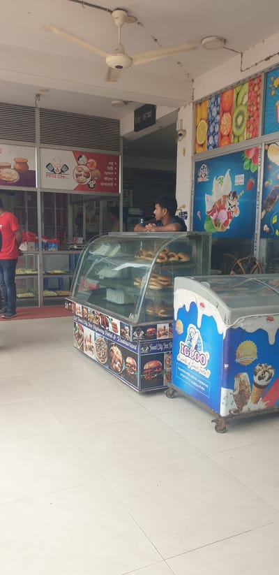 The image shows the exterior of a food establishment, likely a restaurant or cafe, located on the highway in Hatikumrul, Bangladesh. A ceiling fan is visible at the top of the frame, and a sign reading "Sweet fast food" is mounted on the wall. The establishment features a glass display case filled with food items, including pastries and sandwiches. Next to it is a blue and white branded cooler labeled "IGLOO" with images of ice cream. Several people are present, including a man in a red shirt standing near the entrance, and another man leaning on the display counter and looking to the right. The surrounding environment suggests an urban or semi-urban setting with tiled flooring. The overall scene depicts a typical commercial space serving food and beverages.