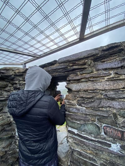 A person is taking a photo through a gap in a rock structure. They are wearing a gray hooded sweatshirt and a black puffer jacket. The rock structure appears to be composed of dark, layered slate. The background visible through the gap and above the rock is a cloudy sky. A metal safety fence is visible to the left of the person. Based on the distinctive slate formations, this image appears to be taken at Dinorwic Quarry, near Llanberis, United Kingdom.