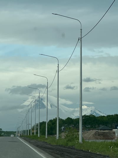 The image depicts a road lined with streetlights leading towards a snow-capped mountain range partially obscured by clouds. The road is paved with a white line marking the lane. On the right side of the road, there is a construction site with piles of dirt and some newly built structures. A line of trees separates the construction site from the mountains. In the distance, on the road, a white car is visible. The sky is cloudy with some patches of blue. The scene appears to be in an outdoor, possibly rural or suburban, setting. The location is identified as Kruetoberegovy, Russia.
