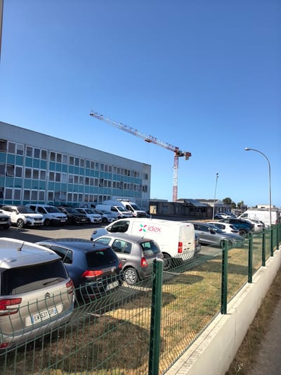 A crane is visible against a clear blue sky, extending over a row of parked cars and a light blue industrial building. The crane's lattice structure is white and red. In the foreground, a green chain-link fence runs along a concrete barrier. Several cars, including a silver Citroën C4 Picasso and a dark blue Volkswagen Golf, are parked behind the fence. White vans and other cars are parked in front of the industrial building, which features numerous windows. The setting appears to be a business or industrial area in Saint-Herblain, France.