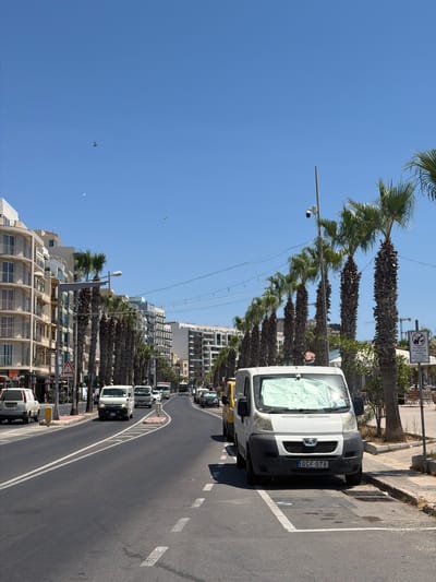 The image shows a street scene in Gżira, Malta, under a clear blue sky. A white van with the license plate GCF 078 is parked on the right side of the road. The van's windshield is covered with a light green sunshade. Palm trees line the street, and several multi-story buildings with balconies are visible on the left. Traffic can be seen in the distance, including cars and a bus. A surveillance camera is mounted on a pole near the palm trees. A sign on the right side of the road displays a pictogram of a person being towed away, with text that reads "NO BEACH UMBRELLAS ON THE STREET". A pigeon is visible flying in the sky.