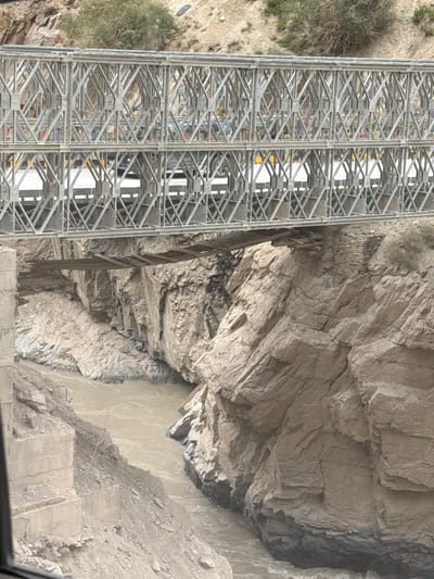 The image depicts a multi-level bridge constructed of a silver-colored metal truss system spanning a narrow, rocky gorge. The upper level of the bridge is a substantial metal structure with cars visible on its surface. Below this, a more rudimentary wooden plank bridge is suspended, appearing weathered and in disrepair, crossing the same gorge. A muddy, fast-flowing river winds through the bottom of the gorge, flanked by steep, textured rock formations. The surrounding environment is arid and mountainous, with sparse vegetation on the slopes. The scene is captured from a perspective that emphasizes the depth of the gorge and the engineering of the bridge, suggesting a remote, possibly challenging terrain.