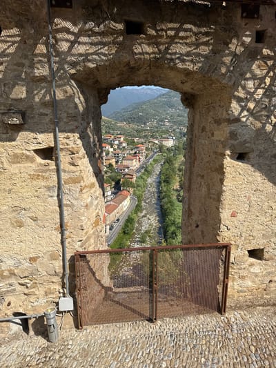 The image captures a view through an arched stone opening in a building, looking out onto the town of Dolceacqua, Italy. The opening reveals a valley with a river, lined by trees and a road with cars. Buildings with terracotta roofs are clustered on the hillsides. The foreground consists of a cobblestone path and a rusted metal grate. The stone walls of the building framing the view are textured and show signs of age. Sunlight casts shadows through a patterned structure above the archway, creating a textured effect on the stone. Electrical conduits and a light fixture are visible on the left side of the stone wall. The scene is bathed in daylight, indicating it was taken during the daytime. There are no people visible in the image.