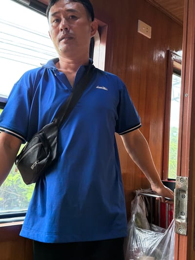 The image shows a man from the chest up, wearing a blue polo shirt with a black fanny pack worn across his chest. The man is looking slightly to his left, with his mouth slightly open. The background is the interior of a train car, with wooden paneling, a window showing greenery outside, and a power outlet on the wall. The man's right arm is bent, and his hand is resting on a metal bar on the right side of the frame. This is likely a train traveling through Hải Vân Ward, Vietnam, known for its scenic coastal railway line.