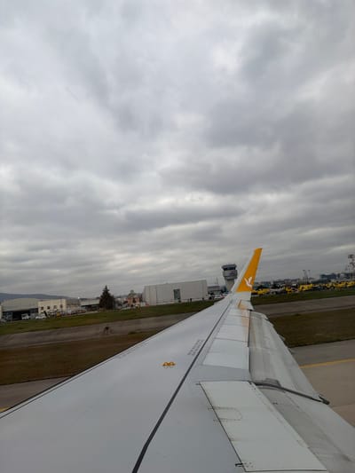 The image depicts an airport scene from the perspective of an aircraft window, showing a portion of the right wing in the foreground. The location is Sabiha Gökçen International Airport (ISG) in Pendik, Istanbul, adjacent to Tuzla, Turkey.

The aircraft wing, predominantly white with a dark gray leading edge and black stripe, extends from the lower right into the mid-ground. The winglet or vertical stabilizer, positioned towards the rear of the wing, is yellow and features the Pegasus Airlines logo (a stylized bird). Visible markings on the wing include text such as "NO STEP" and "DO NOT STEP ON THIS AREA" in black. The wing's flaps and slats appear retracted or partially extended.

Beyond the wing, an airport taxiway or tarmac is visible, characterized by gray asphalt and yellow painted lines. Adjacent to this, a strip of green grass and brown earth separates the immediate ground from airport buildings. In the mid-ground, a multi-story airport control tower stands prominent, gray in color, with a visible "ISG" logo near its top. To the left of the control tower, several industrial-style airport buildings and hangars are present. To the right of the control tower, numerous yellow ground support vehicles are parked in an organized row on the tarmac. A distant cityscape or hills are faintly visible on the horizon behind the airport infrastructure.

The sky is entirely overcast with a dense layer of gray clouds, indicating a cloudy or gloomy day. No discernible individuals or specific human actions are clearly depicted within the scene. The overall impression is one of an aircraft on the ground at an operational airport.