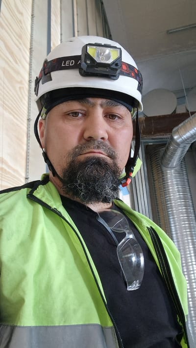 The media file is a portrait of a man wearing a white hard hat with a headlamp. He has a beard and is wearing a green safety vest over a black shirt, with safety glasses tucked into the vest. The background appears to be an indoor industrial or workshop setting, with wooden planks and metallic pipes visible. The lighting suggests it's daytime. The man's expression is neutral. The headlamp on his hat has "LED A" visible as text on the strap.