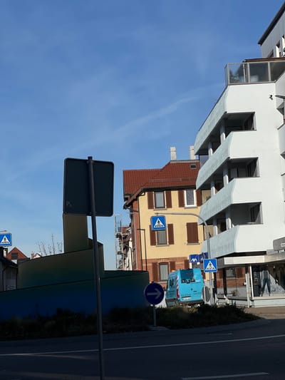 The scene depicts a sunny urban street view in Kernen im Remstal, Germany, under a clear blue sky with a faint contrail. In the immediate foreground, a dark asphalt road surface is visible. A dark grey rectangular traffic sign, blank on the visible rear, stands centrally on a pole. To its right and slightly behind, a circular blue traffic sign with a white right-pointing arrow is present. A low patch of dark green foliage sits on a median between the road and structures. To the left of the central signs, a large, spiraling structure with green and blue surfaces is partially visible. A light blue delivery van, marked "BUG 24/7" on its rear, is parked or positioned behind the foliage in the mid-right. A curved street pole supports two blue square traffic signs depicting a white pedestrian crossing symbol, one positioned near the van and another higher up.
In the background, a series of buildings line the street. On the far left, a multi-story building with a red-tiled roof has scaffolding visible on its left side. Adjacent to it, a three-story building with a yellow facade features multiple windows with dark brown shutters and a red-tiled roof with two white chimneys. To the right, a prominent multi-story white building with a modern design displays curved, cantilevered balconies with light blue undersides and glass railings on its upper levels, including a rooftop terrace. The ground floor of this white building contains a commercial storefront with large display windows, showing a mannequin. No individuals are discernible in the scene.