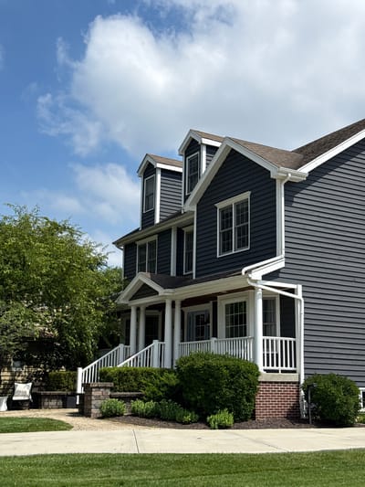 The image shows a two-story dark gray vinyl-sided house with white trim and a brown shingled roof. The house has multiple dormer windows on the upper level and large windows on the first level, some of which are covered by white shutters. A white-railed porch with white columns extends across the front of the house. The house is surrounded by green trees and manicured bushes. A paved walkway leads to the porch. The scene is set on a clear, sunny day with a blue sky and white clouds. The image appears to be taken in a residential area.