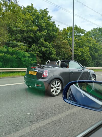 A dark grey Mini Cooper convertible is driving on a road, possibly a motorway, with a woman driving and another passenger in the front seat. The car has a black spoiler on the back and a yellow license plate that reads "YE13 KNH". The road is bordered by a metal guardrail and lush green trees line the side of the road. Overhead, power lines and a tall pole are visible against a cloudy sky. The image is taken from another vehicle, with a blue side mirror visible in the foreground, showing a reflection of the interior of the vehicle and a glimpse of the road ahead. It is difficult to definitively determine the exact location within Dudley, United Kingdom, but the presence of a motorway and extensive greenery suggests an outskirt or suburban area.