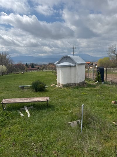 The image depicts a small, white Orthodox church in Gorna Grashtitsa, Bulgaria, situated within a grassy field. The church features a silver cross on its roof. In the foreground, there's a worn wooden bench resting on the grass, and a metal water pump or tap. Beyond the church, there's a view of buildings and mountains under a cloudy sky. A trailer/shed is located behind the church near a fenced area. The overall impression is one of a peaceful, rural churchyard.