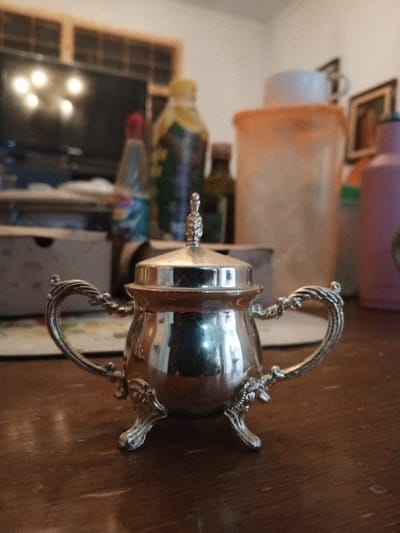 The scene, set in Longmiang, Laos, displays a silver-colored, ornate sugar bowl with a lid positioned prominently in the foreground on a dark wooden table. The sugar bowl features two decorative handles, a finial on its lid, and three stylized legs, presenting a reflective, polished surface. The background, out of focus, reveals an indoor setting with various household items. These include a dark television screen reflecting light sources on the left, and a collection of blurred bottles and containers: a slender bottle with a red cap, a taller bottle with a yellow cap, a dark-colored bottle, a large translucent orange container topped with a white mug, and a pink bottle on the far right. A wooden window frame with horizontal grates is visible above the television, and a framed picture is partially visible on the right wall. No individuals or specific actions are discernible.