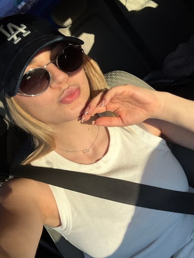 A light-skinned person is lying on their back wearing a white ribbed shirt with a black seatbelt diagonally across their body. The person has a baseball cap with the letters "LA" on the front and round sunglasses. Their left hand is raised with their fingers near their mouth. A delicate silver necklace is visible around their neck. The setting appears to be inside a car. The location in the city of San Jose, United States of America, cannot be determined from the image.