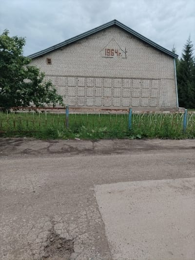 The image shows a long, light-colored brick building with a gabled roof and a dark grey edge. Centered on the upper part of the building, within a triangle of brickwork, are the red painted numbers "1964" followed by a red cross symbol. Below this, the building's facade features a pattern of recessed rectangular shapes arranged in multiple rows. A small, square ventilation opening is visible in the brickwork on the left side of the building. In front of the building, there is a rusty metal fence with decorative elements and blue painted posts, behind which is a grassy area with some weeds. To the left of the building, a green tree with dense foliage is partially visible. The foreground of the image shows a rough asphalt road with a large pothole. The sky is overcast and grey. The scene appears to be outdoors in a rural or semi-urban setting, possibly in Nizhnyaya Maktaama, Russia, given the presence of Cyrillic script on the building. There are no people or animals visible in the image.