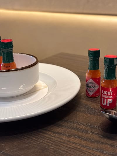 Here is a description of the image:

The photograph shows a tabletop setup that includes a small white bowl nestled on a white plate. Near these dishes are two miniature bottles, one of which is clearly labeled "TABASCO BRAND PEPPER SAUCE" with a volume of 3.7ml (0.12oz). The other bottle has a label stating "LIGHT THINGS UP." These items are positioned on what appears to be a wooden surface. The color palette is neutral, with the browns of the wood, whites of the dishes, and the orange-red tones of the hot sauce. The photograph provides a close-up, detailed view of the items on the table.