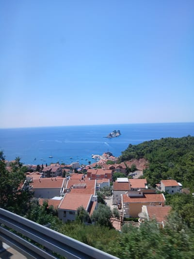 The image captures an elevated view of a coastal town nestled between lush green hills and the azure Adriatic Sea. In the foreground, a metal railing, possibly part of a balcony or overpass, runs horizontally across the bottom of the frame. Beyond the railing, a cluster of buildings with terracotta tile roofs dominates the mid-ground. These structures appear to be residential or commercial, with a mix of architectural styles, some displaying modern features like air conditioning units.
The town extends towards the sea, where numerous boats, including sailboats and smaller vessels, are anchored or in motion. The sea itself is a vibrant blue, stretching out to the horizon under a clear, bright sky. In the distance, a small, rocky island with vegetation adds a picturesque element to the seascape. The hills behind the town are densely covered with trees, indicating a natural, undeveloped landscape surrounding the developed area. The overall impression is one of a serene Mediterranean coastal settlement on a sunny day. While the prompt mentions "Medinski Krš, Montenegro", this specific location is not visually identifiable from the image alone.